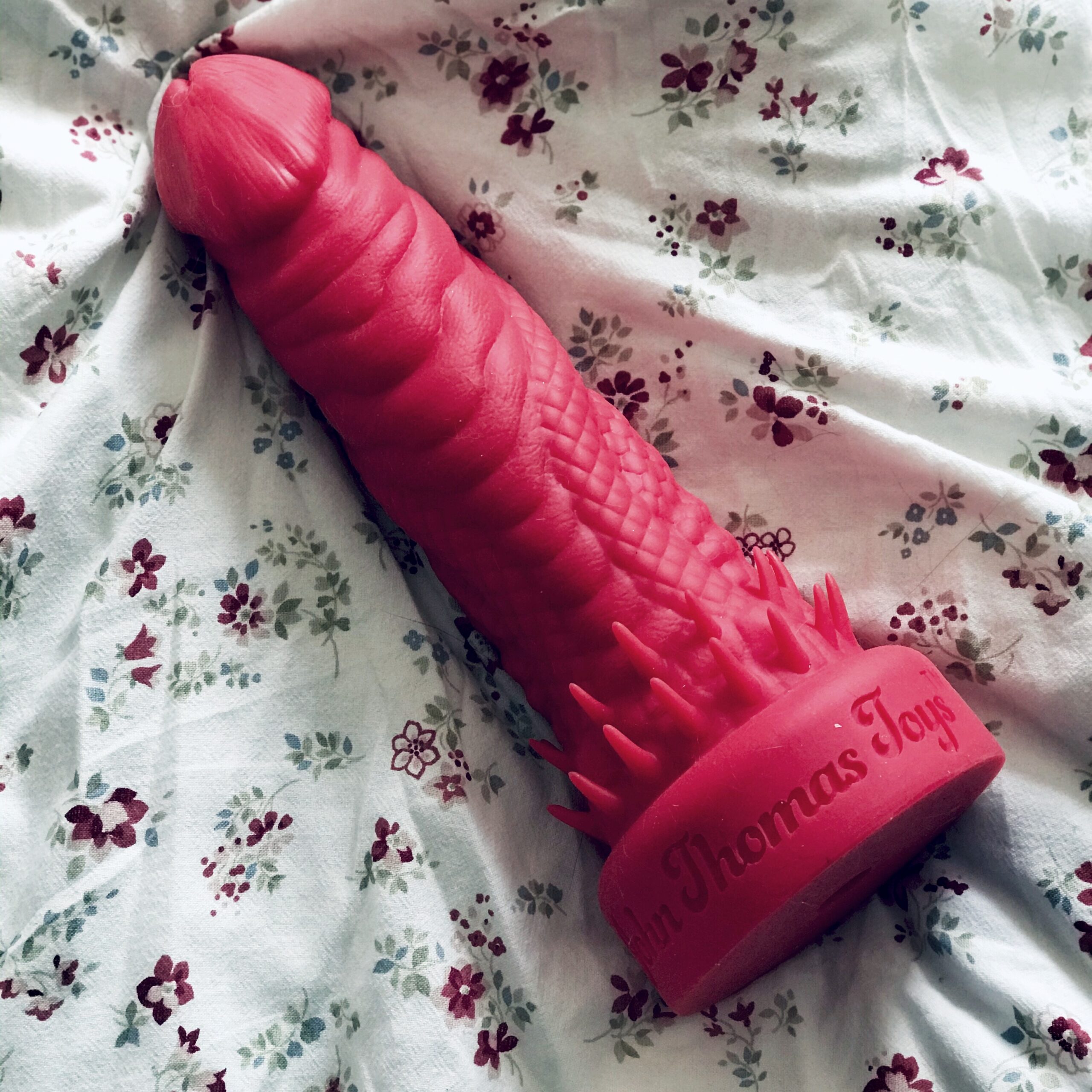 A red Barabarian dildo from John Thomas Toys lays on a duvet covered in an all over floral pattern of red and pink roses. Delicate, feminine floral vs 12" of silicone shaped like and alien dragon penis.